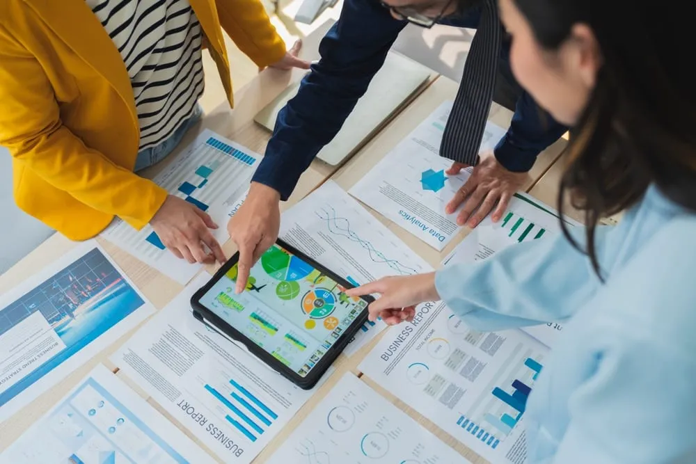 Drie mensen bespreken grafieken en rapporten op tafel en op een tablet